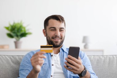 Gülümseyen olgun beyaz adam alışveriş koliği, malların parasını oturma odasında kredi kartı ve akıllı telefonla ödüyor.