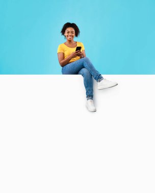 Akıllı telefonlu beyaz pankartta oturan havalı genç siyahi kadın, çevrimiçi servisinizi veya ürününüzü tanıtıyor.