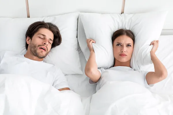 Attractive millennial european guy with stubble sleeps on bed, angry upset wife covers ears with pillow — Stock Photo, Image