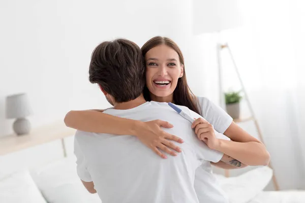 Feliz joven europeo esposa con positivo embarazo prueba abrazos marido en dormitorio interior —  Fotos de Stock