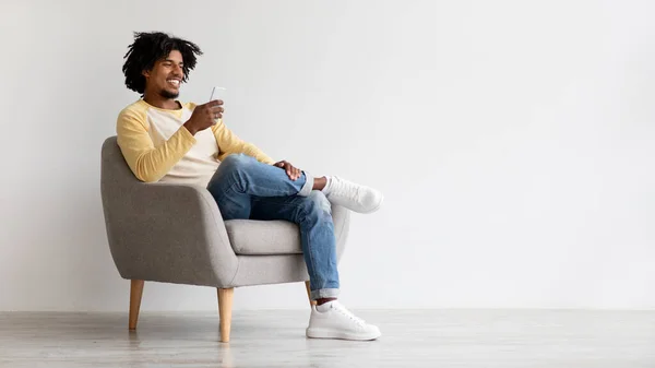 Comunicación móvil. Joven negro chico mensajería en el teléfono inteligente mientras se relaja en sillón —  Fotos de Stock