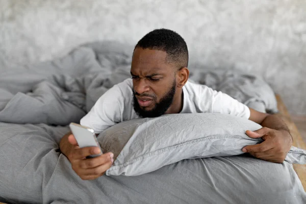 Μπερδεμένος μαύρος που κοιτάει το τηλέφωνο στο κρεβάτι. — Φωτογραφία Αρχείου