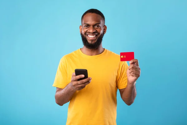 Wesoły młody czarny mężczyzna posiadający kartę kredytową, korzystający z telefonu komórkowego do zakupów online, bankowości lub płatności elektronicznej — Zdjęcie stockowe