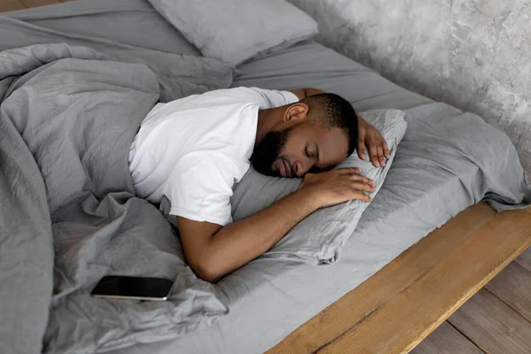 Pemuda Afrika Amerika tidur di tempat tidur dengan ponsel — Stok Foto