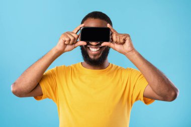 Mutlu siyah adam boş akıllı telefonla gözlerini kapatıyor, web sitesi için modellik yapıyor ya da mavi stüdyo arka planında başvuru yapıyor.