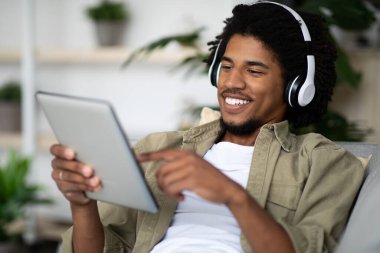 Evde dijital tablet kullanan, kablosuz kulaklıklı mutlu siyah adam.