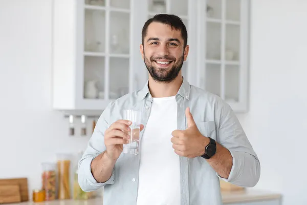 Sorridente bello adulto caucasico uomo tenere il vetro con acqua e mostrando pollice verso l'alto — Foto Stock