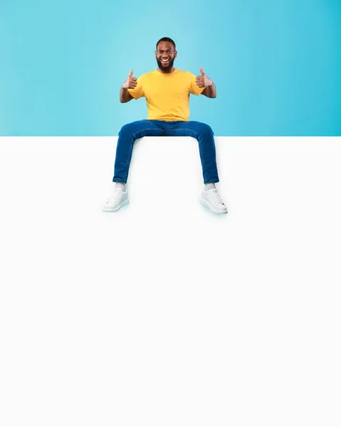 Веселий чорний хлопець сидить на порожньому білому папері банер, показуючи великий палець вгору жест, пропонуючи простір для реклами, макет — стокове фото