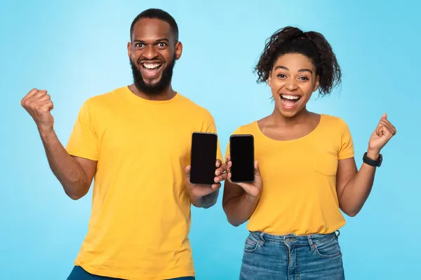 Pasangan kulit hitam yang bersemangat menunjukkan smartphone dengan mockup, gesturing YES, iklan situs baru melalui latar belakang biru — Stok Foto