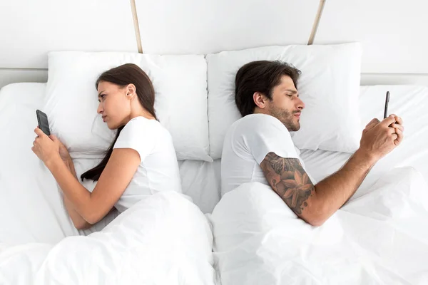 Triste casal milenar europeu conversando em smartphones de volta para trás, deitado na cama no quarto — Fotografia de Stock