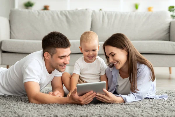 Giovani genitori allegri e bambino bambino che si rilassa con la compressa digitale a casa — Foto Stock