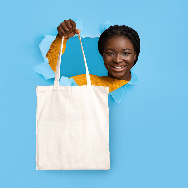 Young black woman holding eco tote bag with mockup for your brand logo design through hole in torn blue paper — Stock Photo, Image