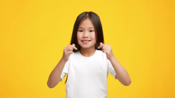 Маленькая корейская девочка показывает сердце символа с пальцами, желтый фон — стоковое видео