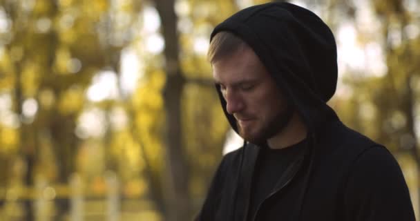 Young caucasian man zipping up his black hoodie, posing at autumn public park and looking aside at empty space — Stock Video