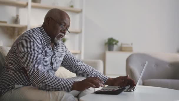 Senior afrikansk amerikansk man redovisning hemma, räkna kostnader och intäkter på miniräknare och bärbar dator, spårning skott — Stockvideo