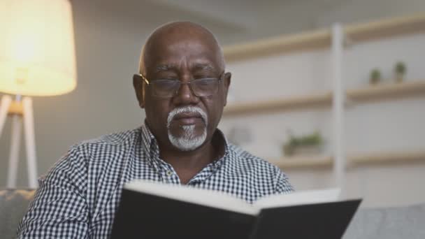 Primer plano del hombre afroamericano mayor concentrado que usa anteojos leyendo un libro interesante, en casa, rastreando disparos — Vídeos de Stock