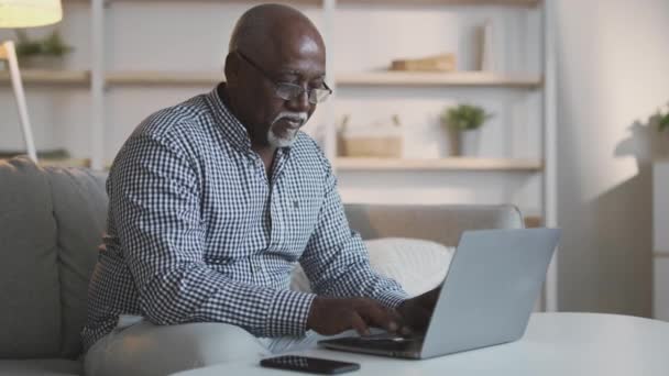 Серьезный пожилой африканский американец фрилансер в очках, печатающий на ноутбуке, работающий из дома онлайн, свободное место — стоковое видео