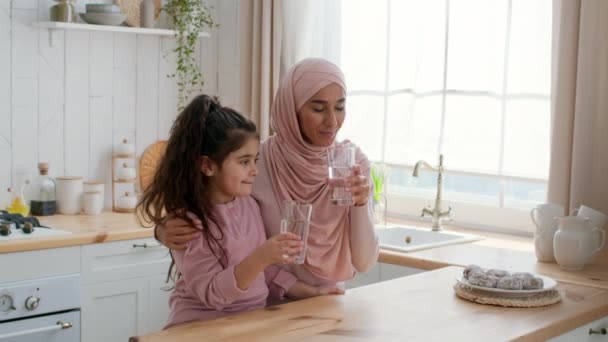 Щаслива мусульманська мати і маленька дочка п'ють воду на кухні — стокове відео