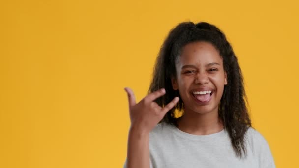 Svart tonåring flicka Gesturing Rock tecken visar tungan, gul bakgrund — Stockvideo