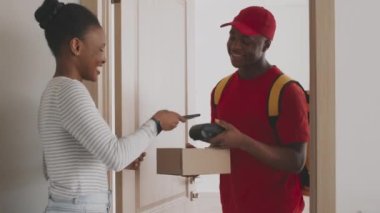 Genç Afro-Amerikalı kadın siparişi akıllı telefonla ödüyor ve posta işçisinden paket kutusu alıyor.