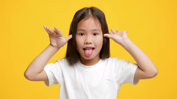 Смешная маленькая кореянка высовывает язык гримасом, желтый фон — стоковое видео