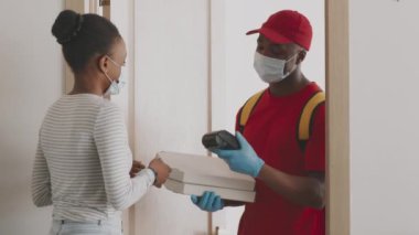 Genç Afrikalı Amerikalı kadın pizza siparişini kuryeden kuryeye akıllı adımlarla ödüyor. İkisi de tıbbi maskeli ve ağır çekimde.