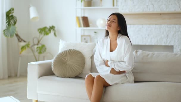 Dor feminina. Jovem mulher asiática sofrendo de dor menstrual, segurando o estômago doloroso no sofá em casa, espaço livre — Vídeo de Stock