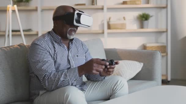 Stárnutí a kybernetický svět. Senior africký Američan nosí VR sluchátka hrát videohry s joystick, sedí doma — Stock video