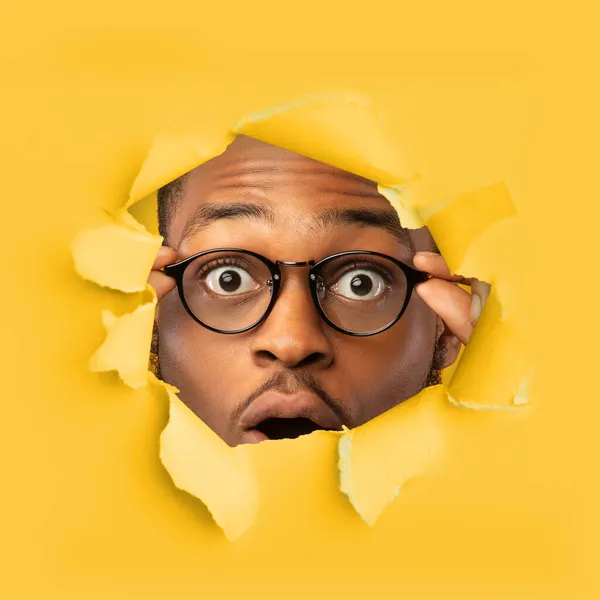 Sorprendido sorprendido milenario hombre negro en gafas boca abierta mira a través de un agujero en papel amarillo, cuadrado — Foto de Stock