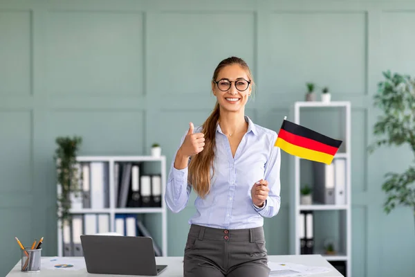 Glückliche Lehrerin mit Deutschland-Fahne, die den Daumen nach oben zeigt, steht in der Nähe des Arbeitsplatzes im Büroinnenraum — Stockfoto