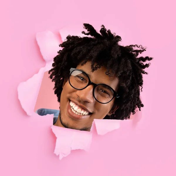 Funny cheerful millennial black man in glasses looks through hole in pink paper, square — Stock Photo, Image