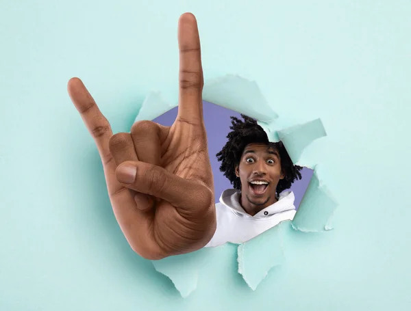 Emocionado adolescente negro mostrando gran gesto de roca a través del agujero en papel rasgado azul claro —  Fotos de Stock