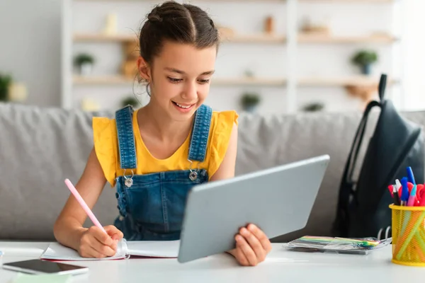 女孩坐在桌旁，用平板电脑，在笔记本上写字 — 图库照片