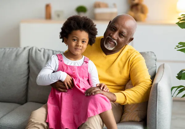 Sorrindo pequena menina afro-americana sentada de joelhos de avô idoso no sofá na sala de estar interior — Fotografia de Stock