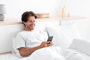 Mutlu Avrupalı milenyum adamı, rahatla, telefonda yazıyor, beyaz yatak odasında yatıyor.