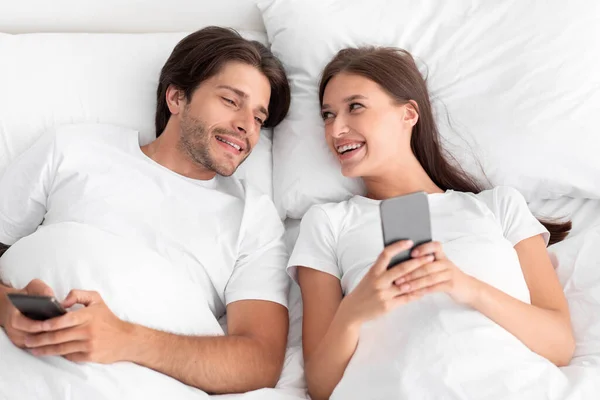 Heureuse femme millénaire caucasienne et mari regardant smartphone, couchée sur le lit dans l'intérieur de la chambre blanche, vue sur le dessus — Photo