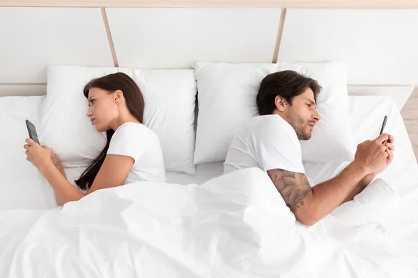 Caucásico millennial macho y hembra yacen en la cama espalda con espalda y charlando en las redes sociales — Foto de Stock
