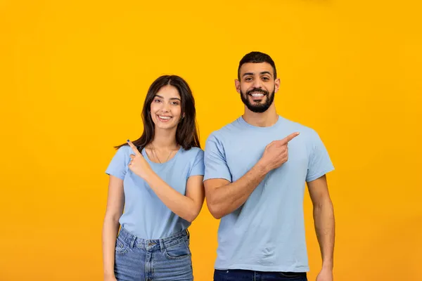 Portrait of excited arab man and woman pointing fingers aside at free space and smiling to camera over yellow background — Stock Photo, Image