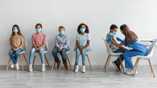 Разнообразные дети получают вакцину против ковида-19, сидя на сером фоне — стоковое фото