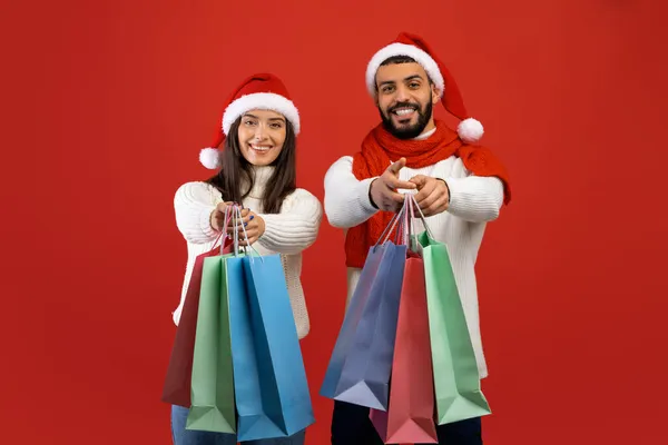 Venta de Navidad. Feliz pareja árabe usando sombreros de Santa y sosteniendo bolsas de compras sobre fondo de estudio rojo — Foto de Stock