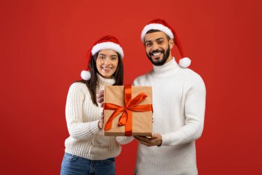 Mutlu Arap eşler Noel hediyesi kutusunu tutuyorlar ve kameraya gülümsüyorlar, kırmızı arka planda Noel Baba şapkaları takıyorlar.