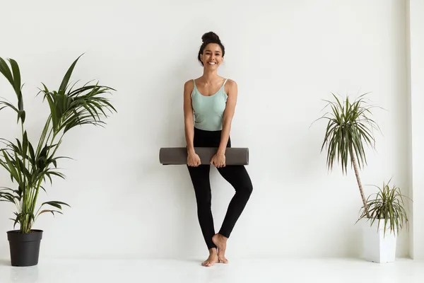 Hermosa mujer joven en ropa deportiva sosteniendo estera de yoga y sonriendo a la cámara —  Fotos de Stock