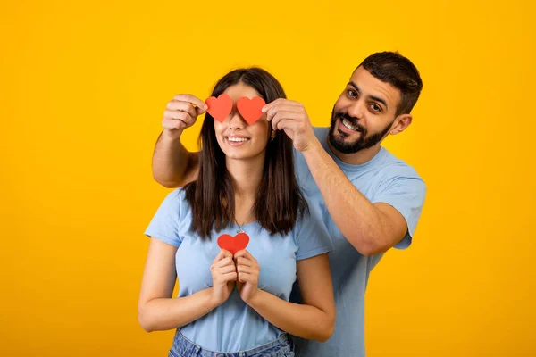 Celebración de San Valentín. amante árabe marido cubriendo esposas ojos con papel corazones, amarillo fondo — Foto de Stock