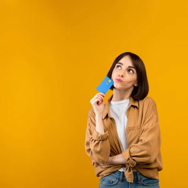 Nieograniczona koncepcja zakupów. Marzycielska młoda kobieta trzymająca kartę kredytową i myśląca co kupić, żółte tło — Zdjęcie stockowe