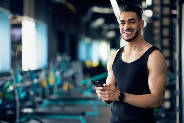 Portré mosolygó jóképű arab fickó használ okostelefon edzés közben a tornaterem — Stock Fotó