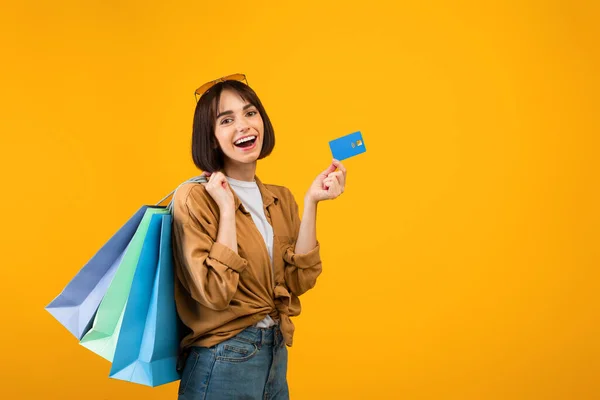 Concetto di Cashback. Donna felice in possesso di carta di credito e borse della spesa luminose, posa su sfondo giallo, spazio libero — Foto Stock