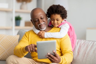 Mutlu siyah kıvırcık kız yaşlı büyükbabaya sarılıyor, tablete bakıyor, video görüşmesi yapıyor, oturma odasında oyun oynuyor.