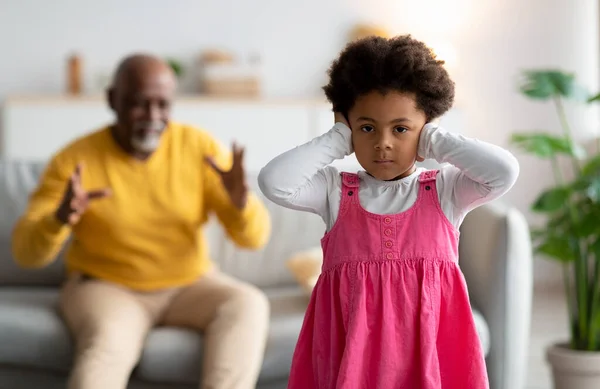 Trauriges schwarzes kleines Mädchen, das ihre Ohren bedeckt. Alter Opa gestikuliert und schwört Enkelin — Stockfoto