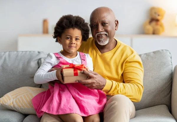 Menina americana africana pequena alegre senta-se de joelhos de homem idoso e prende presente de seu avô — Fotografia de Stock
