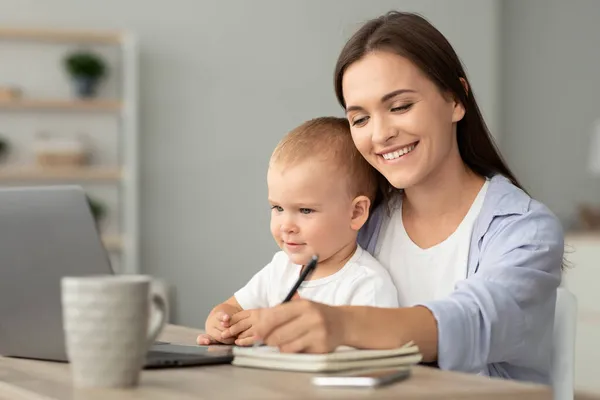 Educação online. Jovem mulher segurando bebê e estudar com laptop em casa — Fotografia de Stock